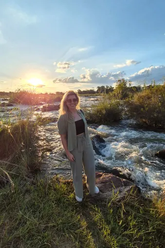 Vrouw staat bij de Popa Falls in Namibië met de rivier en een kleurrijke zonsondergang op de achtergrond.