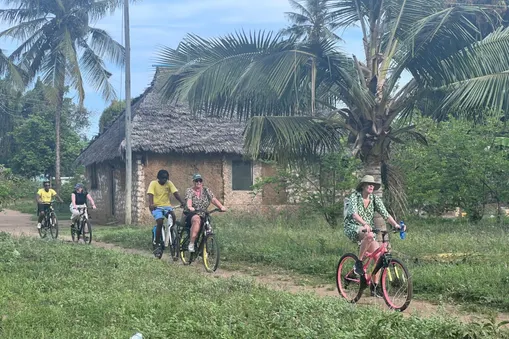 Reizigers fietsen langs een traditioneel Keniaans dorpje met palmbomen tijdens de excursie bij Diani Beach