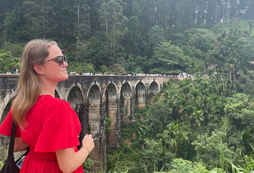 "Vrouw in rode jurk kijkt uit over de beroemde Nine Arches Bridge in Ella, omringd door weelderige jungle en toeristen op de brug."